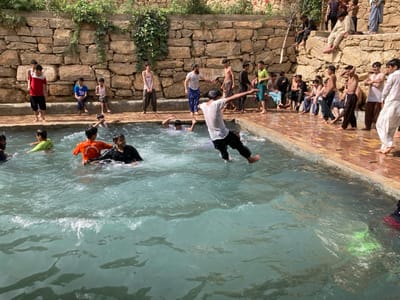 An outdoor scene in Killi Malik Sher Mohammad, Pakistan, depicts a communal swimming area with approximately 25-30 males, ranging from children to young adults. A light blue-green body of water, likely a pool, occupies the lower and central portions of the image. The pool is bordered by reddish-brown tiled paving. In the immediate foreground, a male in a white t-shirt and dark pants is captured mid-air, feet descending into the water, creating a splash. His arms are extended laterally for balance. Several other males are submerged or partially submerged in the water, including one in an orange t-shirt and another in a light green shirt.
In the background, a natural stone retaining wall, partially covered with green foliage, rises above the paved area. Numerous males are positioned on the paved edge, standing, sitting, or observing. Others are seated on higher stone steps and ledges integrated into the wall structure. Clothing varies from shirtless to t-shirts and traditional shalwar kameez-style bottoms, with several individuals appearing wet. The scene is illuminated by bright daylight, suggesting recreational activity in a natural or semi-natural setting.