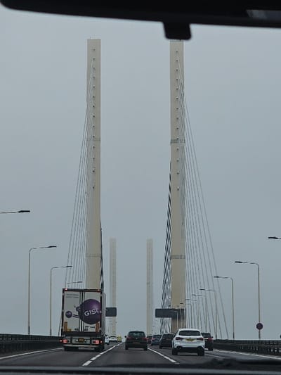 The image, captured from the interior of a vehicle, depicts the Queen Elizabeth II Bridge, part of the Dartford Crossing, located adjacent to South Ockendon, United Kingdom, spanning the River Thames. The scene is under an overcast sky, presenting a uniformly grey ambient light.

Two prominent, light-colored cable-stayed bridge pylons rise vertically in the mid-ground, supporting numerous white tensioning cables that fan downwards towards the bridge deck. Further in the distance, two smaller, uncompleted, or support pylon structures are visible.

The bridge deck carries multiple lanes of vehicular traffic. In the foreground left lane, a white semi-trailer truck with "GIST" branding and a purple circular logo on its rear is observed, displaying illuminated red tail lights. Its license plate is partially visible (e.g., "P7 F441"). In the foreground right lane, a white SUV is present. Immediately behind these, a dark-colored hatchback occupies a middle lane. Several additional vehicles, including a yellow vehicle with flashing lights suggestive of an emergency or recovery service, are visible further ahead in traffic.

Tall streetlights with single luminaires are positioned along both sides of the bridge deck. The uppermost portion of the image shows the dark, curved interior roofline of the photographing vehicle. No individuals are visibly present outside of vehicles.