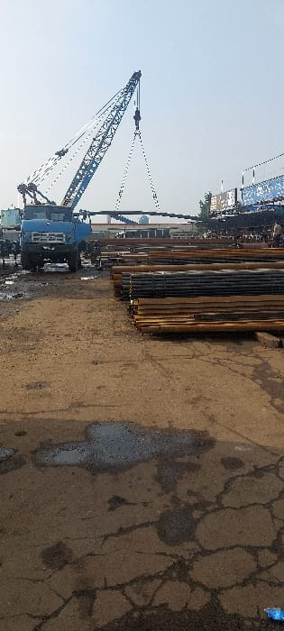 The image depicts an outdoor industrial or construction yard in Nazarbek, Uzbekistan, under a clear, bright sky. A blue truck-mounted crane, positioned on the left side of the frame, is actively lifting a long, dark, horizontal object, suspended by cables from its boom. In the foreground and midground, numerous bundles of metal pipes or cylindrical objects are stacked horizontally on the ground. The ground surface is uneven, composed of dirt and cracked asphalt, with several dark puddles and oil stains visible, indicating wet or muddy conditions in areas. To the left of the crane, indistinct figures of people are present near the truck. In the background, behind the stacks of material and the crane, various industrial structures or buildings are visible, including what appears to be a large, dark structure on the right with some blue signage or coverings. The overall scene suggests ongoing heavy-duty material handling operations.