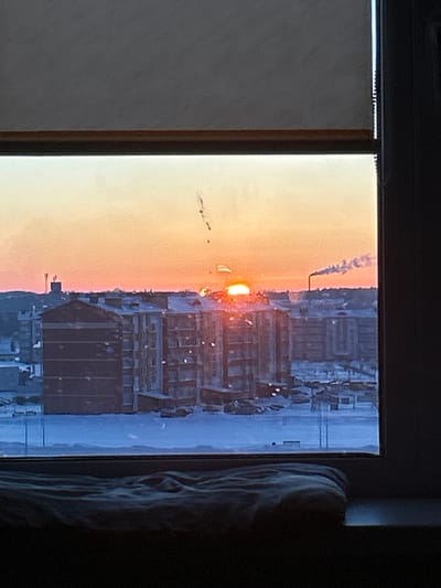 The image displays a snow-covered urban environment in Hlybokaye, Belarus, viewed through a window. A dark gray blind partially covers the top of the frame, and a dark window frame borders the right edge. The scene is illuminated by a low-lying sun, suggesting either sunrise or sunset, which appears as a bright, glowing orange disc above the buildings. The sky exhibits a gradient of intense orange and yellow near the horizon, transitioning to lighter tones and pale blue higher up.
Below the sky, the landscape is blanketed in a thick layer of white snow. In the midground, multiple dark, multi-story residential buildings, likely apartment blocks, are prominent. Their roofs and some external ledges are covered in snow, which reflects the sun's bright orange light. Several parking areas between and around these buildings contain numerous parked vehicles, mostly cars, partially obscured by snow. To the far right, a tall, slender industrial chimney emits a plume of white smoke or steam that drifts across the orange sky. On the far left, a distant communications tower or similar industrial structure is visible. In the immediate foreground, a crumpled dark fabric object, possibly a blanket or cushion, rests on a dark windowsill. No people or discernible interactions are visible within the frame.