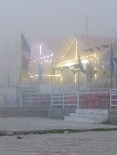 The image depicts an outdoor scene enveloped in thick fog. The primary subjects are a building with a red roof, adorned with festive string lights that glow through the mist. In front of the building, there is a white railing, and beyond that, red chairs and tables are arranged, suggesting a seating area. Several flags are visible, including one with blue, white, and red colors, and other prayer flags strung between poles. A set of white concrete steps leads up towards the building. The ground in the foreground is paved with interlocking bricks, with some patches of grass and scattered stones.

The weather is clearly foggy, obscuring much of the background and creating a soft, diffused light. The time of day is unclear due to the fog, but the illuminated lights suggest it could be evening or early morning. The setting appears to be a public area, possibly a restaurant or cafe, in Marhi, India, given the context. There is no discernible activity taking place; the scene appears static and deserted, contributing to a quiet and somewhat mysterious atmosphere due to the fog. No text is visible in the image.