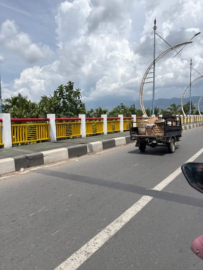 The image shows a three-wheeled vehicle, likely a tuk-tuk or auto-rickshaw, loaded with goods, driving on a road. The vehicle is black and appears to be carrying various items, including what looks like boxes and baskets. The road has white lane markings. To the left of the road, there is a yellow and red striped railing along the side of what appears to be a bridge or elevated walkway. Beyond the railing, there are lush green trees, including palm trees, and a cloudy sky with blue patches visible. In the background, there are large, white, decorative structures that resemble arches or stylized waves, and a hazy mountain range.
The perspective suggests the photo was taken from another vehicle moving in the same direction. A portion of a car's side mirror is visible in the top right corner. The scene appears to be a bright, daytime setting with a mix of clouds and sunshine. The presence of palm trees and the mountainous background could indicate a tropical location. The overall impression is one of daily life and transportation in a possibly coastal or island environment, consistent with Meunasahtutong, Indonesia. No visible text is present in the image.