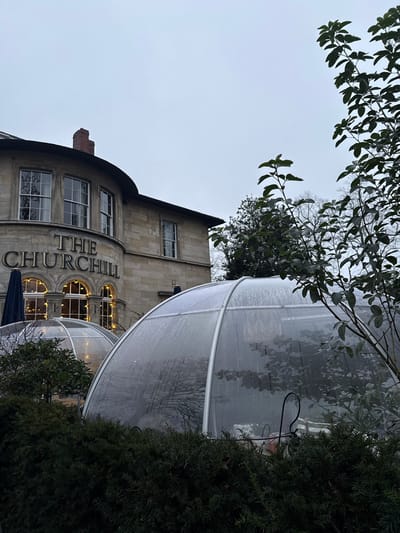 The Churchill building is visible, partially obscured by outdoor dining bubbles. The stone structure has multiple windows and arched details, with "THE CHURCHILL" vertically displayed. The clear, dome-shaped bubbles have metal framing and appear to be situated on a patio. Branches of trees and bushes frame the scene, with an overcast sky in the background, suggesting it may be raining.