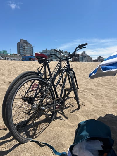 A black bicycle lies on a sandy beach under a bright, clear sky. Buildings are visible in the distance. A blue and white striped beach umbrella partially obscures the view. A dark blue bag sits nearby on the sand. The bike's wheels and frame are casting shadows on the sand.