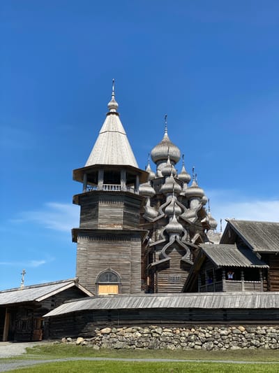 This image shows the Church of the Transfiguration on Kizhi Island, near Boyarshchina, Russia. The UNESCO World Heritage site is a wooden church with numerous domes and a bell tower, all constructed from wood. The church is set against a clear blue sky. In the foreground, a stone wall separates the church grounds from a grassy area. The church features intricate wooden carvings and traditional Russian architecture. The sunlight highlights the weathered wood, showcasing its age and craftsmanship.