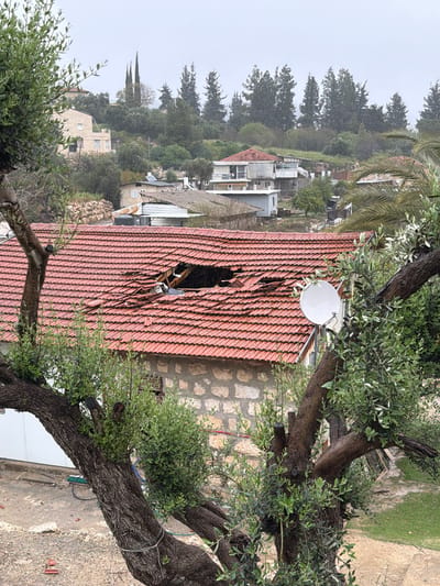 The image depicts a damaged red-tiled roof with a visible hole in it. A white satellite dish is mounted on the roof near the upper right side. Trees with green foliage partially obscure the view. In the background, there is a residential area with houses, trees, and a cloudy sky. The image appears to have been taken in a hilly region, and the environment is likely Mate Yehuda Regional Council, Israel.