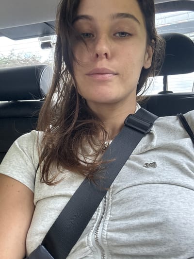 A young woman is shown from the chest up, wearing a light gray zippered hoodie over a light gray t-shirt. A black seatbelt is fastened across her chest. She has long, dark brown hair that is slightly messy and falls around her face and shoulders. Her eyes are open, and she is looking towards the camera with a neutral expression. Her lips are slightly parted. A small, silver charm, possibly a small animal, is attached to the left side of her hoodie. The background appears to be the interior of a vehicle, with the headrest of a dark seat visible behind her. Light streams in from windows on the left side of the image, suggesting it is daytime, possibly overcast. The overall impression is casual and perhaps a moment captured while traveling.