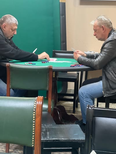 The image displays two adult males seated opposite each other at a green-felt card table, engaged in a card game. The scene is located indoors, within the city of Smederevska Palanka Municipality, Serbia.

The male on the left, with short graying hair, is observed in profile, wearing a black long-sleeved top and blue jeans. He is leaning forward, his right hand positioned near a stack of red poker chips and several playing cards on the green table surface. A collection of blue and red poker chips is arrayed in front of him.

The male on the right, also in profile, exhibits short light-gray hair and is dressed in a black leather jacket and light blue jeans. He holds a hand of playing cards in both hands, directing his gaze downward at them. A distinct arrangement of red and blue poker chips is present on the table before him.

The central object is an oval-shaped card table featuring a green felt playing surface and a dark-toned trim and base. Both players are seated on dark, simple-design chairs with backrests. In the foreground, two unoccupied green upholstered chairs with dark wooden frames and brass stud detailing are partially visible.

The immediate background to the left consists of a solid green wall or panel, while the background to the right is a light beige-colored wall. The floor beneath the table is covered with a patterned carpet. The ambient lighting is uniform, consistent with an interior setting.