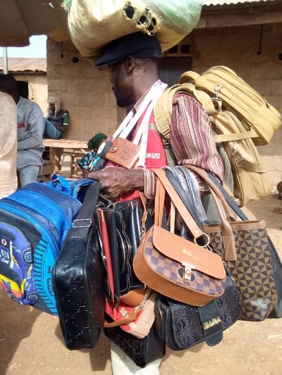 A vendor, possibly African, carries a large sack on his head and numerous bags of various styles and sizes. He has backpacks, shoulder bags, and handbags draped all over him. One brown wallet labeled "GUCCI" hangs around his neck. He also has many metal bracelets on his left hand. In the background are people sitting on benches outside a weathered building.
