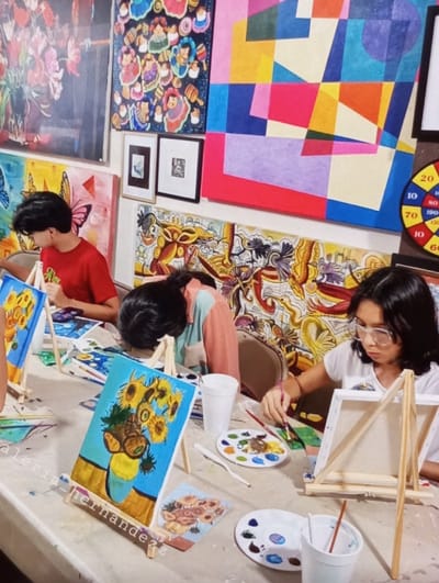 An indoor art workshop in Aktash, Uzbekistan, is depicted, showing three individuals engaged in painting activities at a light-colored table. On the right, a young female with dark hair and clear-framed glasses, wearing a white shirt, meticulously paints on a small canvas affixed to a wooden easel. To her left, a person with dark hair, wearing a striped peach and mint green shirt, is leaned forward over the table, with a painting of sunflowers on an easel in front of them. Further to the left, partially visible, another individual in a red shirt is also painting on an easel.
The table is cluttered with art supplies, including multiple white plastic palettes containing various paint colors, paintbrushes, white plastic cups (some holding brushes), and a small printed reference image of sunflowers. The walls are extensively decorated with numerous artworks: a large framed floral painting (pink flowers in a vase) in the upper left, a vibrant painting of stylized figures in the upper center-left, a large abstract geometric painting in bright colors (pink, blue, yellow, red, purple, orange) in the upper center-right, and two smaller framed artworks below it. A long, horizontal mural with intricate, colorful organic designs spans the mid-level wall behind the participants. A circular dartboard-like object with numerical scores is partially visible on the far right wall. A large painting of butterflies in blue and pink tones is visible on the lower left wall.