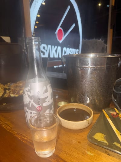 An indoor scene at what appears to be a restaurant location in El Nido, Philippines is captured. The image contains a nearly empty bottle of sake oriented horizontally, a full sake cup, a small bowl of sauce (likely soy sauce), a metallic pot covered in condensation, and a plate with wasabi, all resting on a wooden surface. In the background is a blurred light-up sign with the words "SAKA CASTLE" visible.