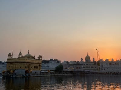 The image captures the Golden Temple (Harmandir Sahib) in Amritsar, India, during sunset. The iconic golden structure, situated in the middle of a reflective pool, is illuminated by the warm hues of the setting sun. The sky transitions from a soft blue at the top to a vibrant orange and yellow near the horizon. Surrounding buildings, also featuring domed architecture, are visible in the background. Several people can be seen gathered on the walkway leading to the temple and along its periphery, with their reflections shimmering in the water. The scene conveys a sense of tranquility and spiritual reverence.