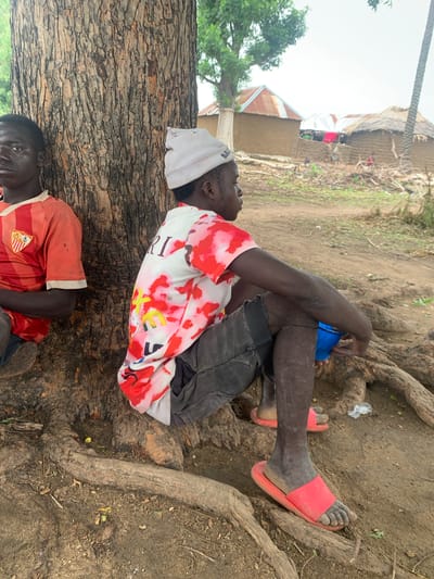 In Dull A, Nigeria, two young men are pictured sitting under a large tree. The man on the left, wearing an orange and red striped t-shirt, leans against the tree trunk. To his right, another young man, wearing a white beanie and a t-shirt with a red and white tie-dye pattern, is seated with his knees bent. He holds a blue bowl in his hand and is wearing dark shorts and red sandals. In the background, several mud-brick houses with corrugated iron or thatched roofs are visible, suggesting a rural or semi-urban environment. The ground is dirt and scattered with dry leaves and debris.
