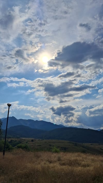 The image depicts a natural landscape scene in Dobrotino, Bulgaria, under a bright, partly cloudy sky. The sun is visible, peeking through scattered clouds, suggesting it is daytime, likely late morning or early afternoon. The scene is split into three distinct parts: the sky, the mountains, and the foreground.

The sky is a vibrant blue, filled with a mix of white and gray clouds. Some clouds are thin and wispy, while others are more dense and dramatic, creating interesting shadows and highlights.

In the mid-ground, a series of mountains dominate the view, receding into the distance. The mountains are covered in lush, dark green vegetation, and their slopes show evidence of terraced farming or natural contours. The topmost peaks are bathed in sunlight, contrasting with the shadowed valleys.

The foreground is divided between a field of dry, golden grass on the right and a dark, silhouetted object on the left. The grass appears to be tall and somewhat dry, possibly indicative of a late summer or early autumn setting. The silhouetted object is a vertical pole with a light fixture at the top, a man-made element in the natural environment. This pole is positioned in the lower left quadrant of the image.

The overall impression is one of a serene and picturesque rural landscape, with the sun casting a warm glow on the scene. There is no visible text in the image.
