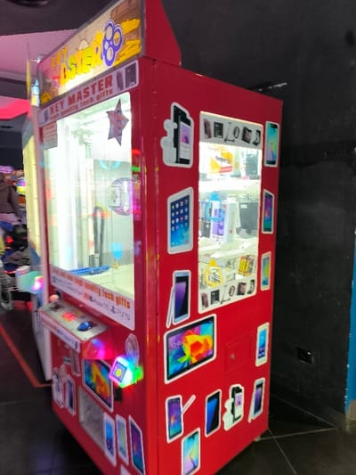 The image displays a vibrant red arcade game machine, identified as a "Key Master," situated in an indoor entertainment venue in Agios Dimitrios, Greece. The machine is tall and rectangular, with a transparent front casing that reveals an array of high-quality tech prizes, including boxed smartphones, tablets, smartwatches, and headphones. A mechanical claw or key mechanism, central to the game, is visible within the prize area.

The machine's exterior is adorned with numerous white silhouette decals depicting various modern electronic devices, reinforcing the theme of the prizes. At the player's level, a control panel featuring illuminated buttons and a joystick is visible, though no one is actively playing. The internal lighting of the machine is bright, contrasting with the dimmer background.

The setting appears to be a public entertainment space, possibly an arcade or family entertainment center, given the dark walls and floor visible in the background. A blurred figure of a person can be partially seen to the left of the machine, suggesting other patrons are present. A dark wall and a blue-paneled column are visible to the right. The artificial lighting suggests an indoor setting, and no specific time of day is discernible.

Visible text on the machine includes "KEY MASTER" prominently displayed on both its top marquee and a smaller panel. Below this, the slogan "win high quality tech gifts" is clear. Further down, examples of the prizes are listed: "Apple Watch," "iPhone16," and "PS5."