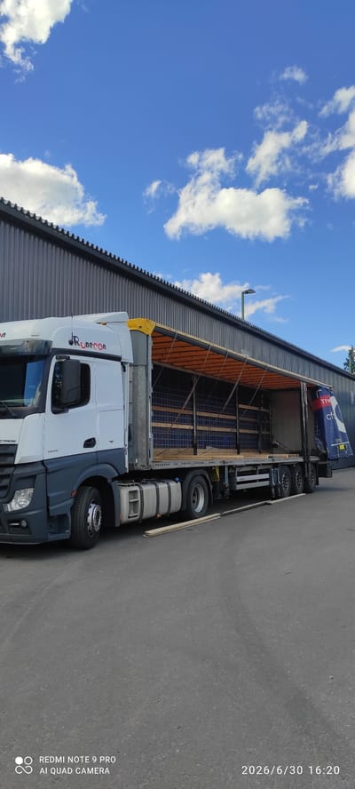This is a landscape-oriented image, photographed outdoors on a sunny day with some white clouds against a bright blue sky. The primary subject is a white semi-truck with "RuBecon" and "ACTROS" text visible on its side. The truck appears to be parked next to a corrugated metal building with an orange canopy extending over a loading area. The truck's trailer is open, showing wooden supports and blue netting inside.
The timestamp "2026/6/30 16:20" is visible in the upper right corner of the image, suggesting the photo was taken in the late afternoon. The text "REDMI NOTE 9 PRO AI QUAD CAMERA" is also present in the lower right corner, indicating the device used for capture.
The overall scene depicts a commercial setting, likely a logistics or distribution center, with a truck being loaded or unloaded. The weather appears to be pleasant, suitable for outdoor work. No people are visible in the frame, and the focus is on the truck and its surroundings. The location is identified as Hankasalmi, Finland.