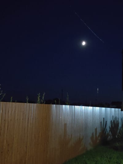 A night-time, wide-angle photograph depicts a dark blue sky over the city of Shkurupiivka, Ukraine. In the upper right quadrant of the sky, a faint, light-colored streak, possibly a contrail or meteor trail, diagonally traverses the frame. Slightly below and to the left of this streak, a bright, half-illuminated moon is visible. A wooden fence, running horizontally across the lower half of the image, is illuminated from above by a light source not visible in the frame, casting shadows of sparse vegetation on its surface. The fence is constructed of vertical wooden planks and appears weathered. In the foreground, grass is visible, dimly lit.