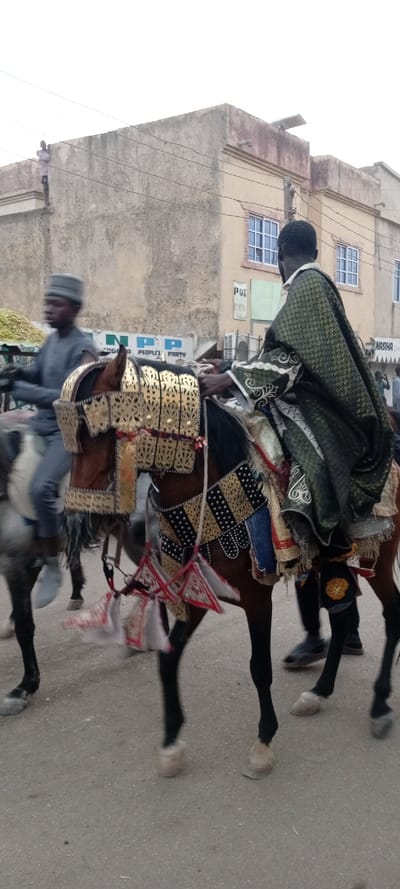Two men on horseback are shown. The horse in the foreground is decorated with gold and black armor, and the rider wears a green robe. Behind them is another horse and rider. A building with windows is in the background. On top of the building, a person is visible. A sign saying "NPP PEOPLE'S PARTY" is also visible.