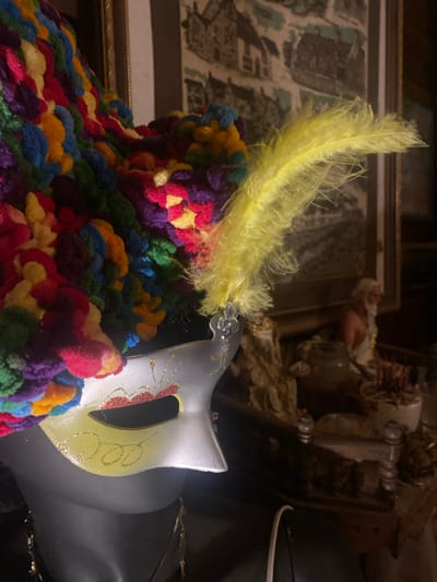 A photograph shows a silver mask with a yellow feather lying on top of a colorful crocheted item. The mask has glittery gold and red designs. In the background, there is an unclear picture in a golden frame and other decorative items. The location is difficult to determine from this image, and there is no evidence to suggest it is in Baan Don Sai, Thailand.