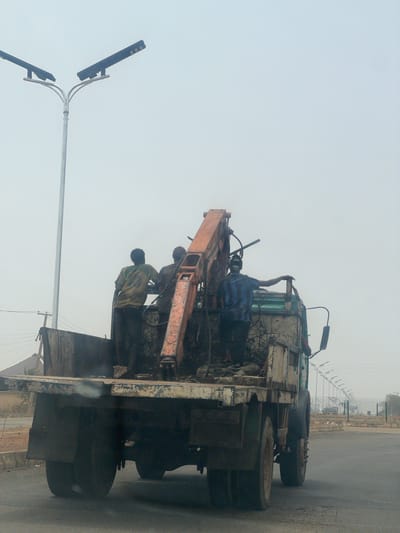 On a road in Haifam, Nigeria, three people are working on a truck with a crane attached. One person is in the truck's cab, and two are standing on the flatbed, working on the crane. The truck is dirty and appears to be old. There is a streetlight to the left of the truck. The sky is overcast, and the road is paved. Fences line the road in the background.