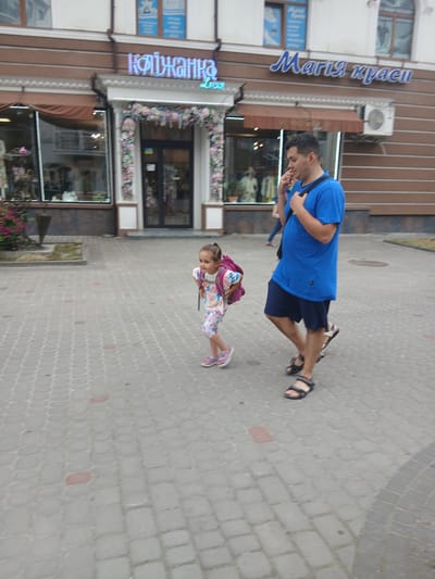 The image captures a street scene in Truskavets, Ukraine. Two people are visible: a man wearing a blue t-shirt and dark shorts, and a young girl in a colorful dress and pink sandals. The man is walking, his head turned downwards as if looking at something. The girl, with a backpack, is also walking. They appear to be on a paved sidewalk in front of a building with storefronts.

One storefront has a blue neon sign with Cyrillic text that reads "Марія краси" (Maria krasy), which translates to "Maria Beauty". Another sign, also in blue neon, reads "Квіжанка" (Kvizhanka), which could be a shop name. The building has a modern facade with windows and vertical brown paneling. Flowers are visible decorating the area above the storefront windows. An air conditioning unit is mounted on the building.

The scene suggests daytime, with ambient light illuminating the area. The ground is paved with cobblestones. The overall atmosphere appears to be one of casual outdoor activity on a street or promenade.