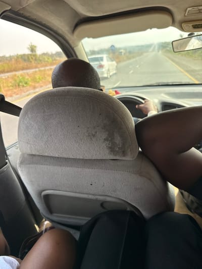 An interior view from a vehicle in Mayo, Nigeria, shows the back of a bald male driver's head and a gray fabric driver's seat, exhibiting multiple dark stains and wear. The driver's right hand grips the steering wheel, a light-colored watch on his wrist. To the right of the driver, the dark-skinned legs of a front passenger, clad in dark attire, are visible. To the left, the light-clad legs of another rear passenger are partially visible. Through the windshield, a two-lane asphalt road with a yellow median stripe extends into a hazy distance. A white sedan proceeds ahead and to the left. A blue circular road sign is visible further along the road. To the left, a railway track runs parallel to the road. Roadside vegetation appears dry and sparse under a bright, overcast sky.