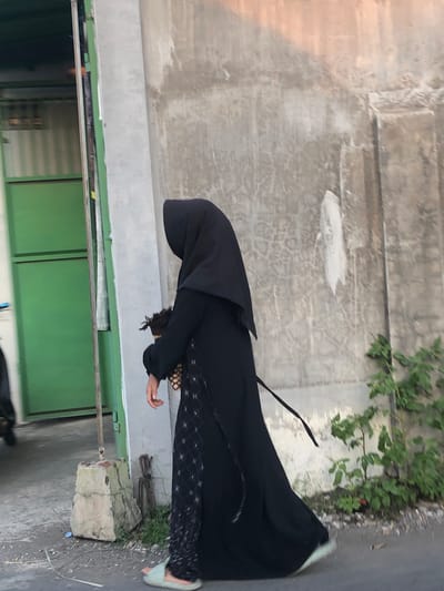 In Glagah Arum, Indonesia, a person wearing a black abaya and hijab walks on a paved surface next to a rough concrete wall. They are carrying a small object in their right hand. To the left of the person, a green metal door is visible, and to the right, a small bush with green leaves grows near the wall. The ground appears to be asphalt or concrete, with some debris and a concrete structure. The lighting suggests it is daytime, with some shadows present.