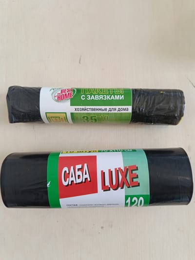 The image shows two rolls of black trash bags on a light-colored, textured surface. The top roll has a label with green and white elements. The text on the label includes "BEST HOME" and "ПАКЕТЫ С ЗАВЯЗКАМИ" (Bags with ties). Below that, it says "хозяйственные для дома" (household for home) and "35 10 шт." (35 10 pcs). There is also a small graphic showing dimensions.
The bottom roll has a label with green and red elements. The text includes "САБА LUXE" (Saba Luxe) and "70 X 110 CM". It also states "10 ШТУК" (10 pieces) and "120" which likely refers to the capacity in liters. The word "СОСТАВ: полиэтилен высокого давления, суперконцентрат полимерный, черный" (Composition: high-pressure polyethylene, polymer concentrate, black) is also visible.
The image is a close-up of the products and does not contain any information about the city of Berdiansk, Ukraine, or any people.