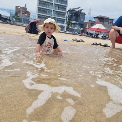 A young child sits in shallow water at a sandy beach, wearing a white sun hat with red polka dots and a black and white raglan t-shirt with a graphic print. The child is looking to the left with a curious expression. The water is clear with small waves and foam washing over the child's legs and hands. The background shows a modern cityscape with tall buildings, some with balconies and awnings. There are umbrellas, beach tents, and several pairs of flip-flops scattered on the sand. The overall scene suggests a pleasant day at the beach in Gangneung-si, South Korea.