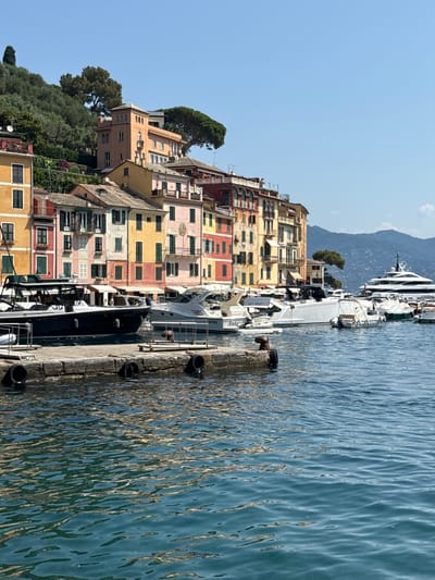 The image depicts a sun-drenched scene in Portofino, Italy, with vibrantly colored buildings lining a hillside overlooking a tranquil harbor. The buildings, painted in shades of yellow, orange, pink, and red, showcase traditional architecture with numerous windows and shutters. Lush green trees and vegetation cascade down the slope towards the water.
Several boats and yachts are moored along a stone pier in the foreground. The water is a deep blue, reflecting the bright sunlight and creating rippling patterns on its surface. A few people are visible on the pier and on one of the boats, suggesting activity within the harbor. The overall atmosphere is one of a peaceful summer day, with clear blue skies and calm seas. The sun appears high in the sky, indicating it is likely midday. The architecture and the picturesque harbor setting are strong cues to the location being Portofino, a well-known Italian Riviera town.