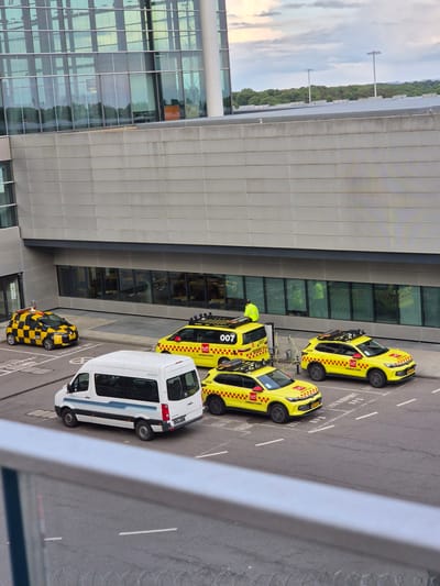 The image shows a parking area outside a modern building, likely an airport terminal. The location is Niederanven, Luxembourg, as indicated by "LUX AIRPOO" on one of the vehicles. There are several vehicles parked in designated parking spots. A white van with the license plate "LG 6725 P1" is visible. There are several yellow cars with "OPERATIONS" and "LUX" on them, and at least two of them have the number "007" and "008" respectively. One of the yellow cars is plugged into a charging station. A person wearing a yellow safety vest is standing near one of the cars. The rest of the parking lot is empty and leads to a modern building with lots of windows and reflective paneling.