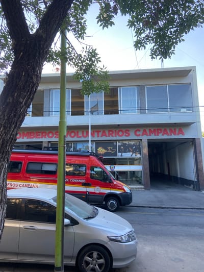 A three-story building displays "Bomberos Voluntarios Campana" vertically in red letters. A red and white fire engine stands parked in front of the building, partially obscured by a white sedan. A tree trunk and leafy branches occupy the foreground. The building has large windows on each floor, and the entrance appears to have images of firefighters. The scene is bathed in daylight.
