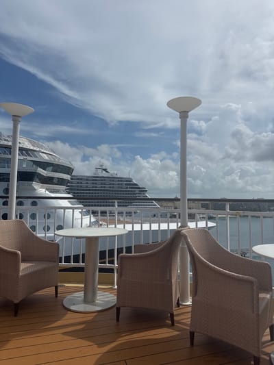 This image captures an outdoor deck, likely of a cruise ship, docked in the city of Nassau, Bahamas. The foreground features a light brown wooden deck floor with prominent shadows cast by sunlight. Two sets of outdoor patio furniture are visible: a round white table with a central pedestal base and two light brown, wicker-style armchairs positioned around it. A similar table is partially visible on the right. Two tall, white light poles with flat, disc-shaped luminaires top them stand symmetrically.

Beyond a white metal railing, the midground is dominated by the massive white hulls and superstructures of two large cruise ships. The ship on the left, closer to the viewer, displays multiple decks with windows and balconies. A second, larger cruise ship is visible behind and to the right of the first, its upper decks and extensive rows of cabins prominent.

In the background, beyond the docked ships, a body of water is visible, indicative of a harbor or port. Distant buildings and structures on the shoreline of Nassau are partially obscured by the ships and the hazy horizon. The sky is partly cloudy with significant white cumulus clouds against a blue sky, indicating daylight hours. No individuals are present in the scene.