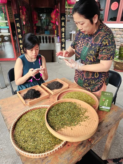 The image depicts a scene in Xihu District, China, with two women in front of a traditional building. In the foreground, a wooden table displays several bamboo trays filled with different types of dried tea leaves. One tray contains a pile of dark tea, while two other trays are filled with lighter green tea leaves. To the right, a green tin can with Chinese text is also on the table.
The woman on the left is seated and wearing a dark teal sleeveless dress with a pink and black beaded necklace. She is holding a white mobile phone and looking down at it. The woman on the right is standing and wearing a short-sleeved, patterned dress with a paisley-like design in shades of purple, green, and orange. She is also holding a white mobile phone and appears to be interacting with it. Her hands are covered with transparent plastic gloves, suggesting she may be handling the tea.
The background shows elements of traditional Chinese architecture, with red lanterns and calligraphy scrolls visible on the building's facade. The overall setting appears to be an outdoor tea stall or shop in a picturesque location.