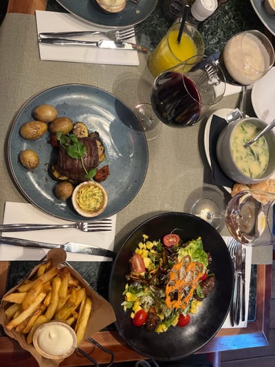 The image displays a dining table set for a meal at an establishment in Baarle-Nassau, Netherlands. The table surface consists of a light green textile runner, a section with a dark green marble pattern, and a dark wooden edge. No individuals are visible.
A blue-grey ceramic plate holds a seared steak, garnished with green shoots, accompanied by roasted baby potatoes and mixed roasted vegetables (zucchini, tomatoes), with a separate small bamboo-like container of herb butter. A place setting with a fork and knife rests on a white napkin next to this plate.
A black ceramic bowl contains a salad featuring mixed greens, diced mango, cherry tomatoes, dark protein pieces, and an orange, intricate, crispy garnish, all dressed with an orange sauce. Below this, a metal basket holds golden-brown french fries and a small cup of white mayonnaise.
On the right side of the table, a grey ceramic bowl presents a light green, creamy soup garnished with chives, with a spoon inside. A basket containing bread rolls is partially visible nearby. Beverages include a glass of red wine, a glass of orange juice with a black straw, and a glass holding a creamy, light-colored beverage. A salt and pepper grinder set is positioned on the table. Another partially visible blue-grey plate at the top holds a small pastry or tart.