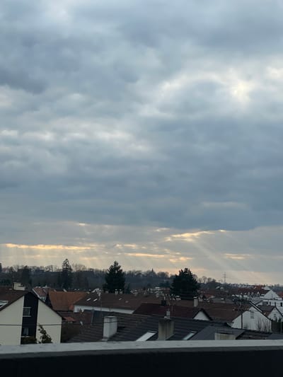An elevated view captures a residential area in Senden, Germany, under a heavily overcast sky. The upper two-thirds of the image are filled with layers of grey clouds, from which multiple distinct crepuscular rays of sunlight descend diagonally towards the horizon, particularly prominent in the mid-left and mid-right. The horizon line reveals a dense band of mostly bare deciduous trees interspersed with several tall, dark evergreen coniferous trees. A distant, dark church spire with a pointed roof is visible above the tree line on the far left. Towards the right, a high-voltage power line tower with multiple cross arms stands against the backdrop of the trees. The lower third of the image is dominated by the rooftops of numerous residential buildings, characterized by a mix of dark grey and reddish-brown tiled roofs. Various chimney structures are present, and some roofs feature skylights and small solar panel arrays. A prominent multi-story house, painted white with dark trim and a steeply pitched dark roof, is visible in the left foreground, partially obscured by a dark, flat railing or parapet running along the very bottom edge of the frame. No human figures or discernible actions are present.