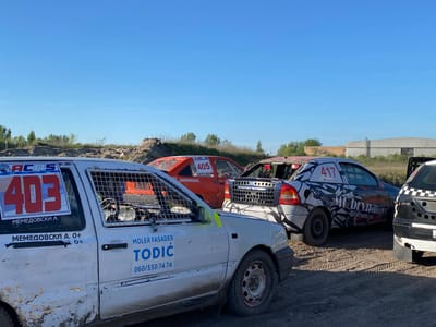 The image shows a dirt track race scene in Kanjiza Municipality, Serbia. Four modified cars are in the foreground:

1.  A white car with the number "403," cage bars over the windows, and a "TODIC" advertisement.
2.  An orange car with the number "405" and the text "HOONIGAN."
3.  A car with a white and black paint scheme, the number "417," and the text "MC bryna."
4.  A black and white car.

The dirt track is visible, and in the background are trees, a dirt hill, and some industrial buildings. The sky is clear and blue.