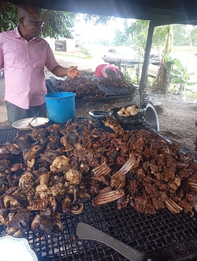 The image depicts an outdoor grilling operation in Assai Sabongida, Nigeria. In the left foreground, an adult male, dark-skinned, wearing a light pink collared shirt, dark trousers, and glasses, extends his right hand towards a substantial accumulation of grilled meat. A white plate with residue is visible near his left side, and a blue plastic bucket rests behind him.
The central feature is a large volume of mixed grilled meats, including ribs, organs, and various other cuts, charred to a dark reddish-brown, distributed across multiple black wire grates over a heat source. A large metal spatula lies on the foreground grill. To the right, a dark bowl holds lighter-colored, possibly fried dough items.
In the background, a second individual, partially obscured by the structure, tends to an additional grill with more meat, wearing a pink garment. The scene is set under a rudimentary, dark-roofed shelter supported by wooden poles. The ground is dirt. Beyond the immediate setup, a dirt road is visible with parked vehicles, including a motorcycle and cars, along with a distant light-colored building and dense green vegetation.