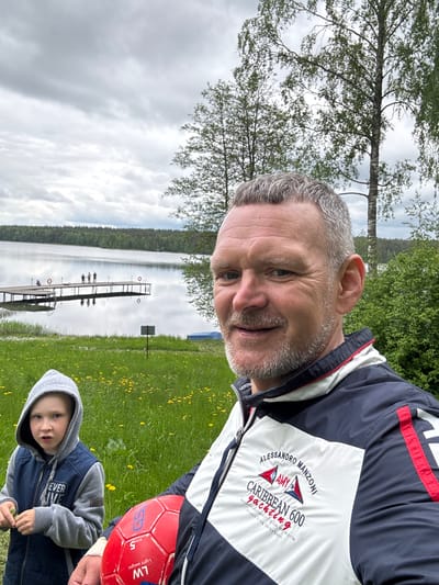 A man and a young child stand in a grassy field near a lake with a wooden pier. The man is wearing a jacket with the words "ALESSANDRO MANZONI," "CARIBBEAN 600 yachting," and the coordinates "Bout Warrior Latst Positio 017 30.665N 06 46.817W" on it. The child is wearing a hooded jacket. A red soccer ball is visible, marked with "GS" and "LW" for "Light weight." The location, based on the jacket's coordinates, is near Aleksandrovka, Russia. The sky is overcast, and trees line the opposite shore of the lake.