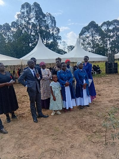 The image depicts an outdoor gathering in Butali Chegulo ward, Kenya, under a partially cloudy sky. In the foreground, a man in a dark suit, white shirt, and blue tie stands, holding a black microphone in his right hand, gesturing slightly. To his left, a woman in a black dress holds a white piece of paper. To his right, a young girl in a light green dress and white sneakers stands facing forward. Behind them, a group of approximately eight women are positioned. Four women wear matching long-sleeved blue tops with white collars and long white skirts. One wears glasses. Other women wear various patterned dresses and head coverings. In the mid-ground, two large white event tents with pointed tops are visible. Under one tent, distant figures are seated. To the far right, a stack of black sound speakers is present. The background is dominated by a dense line of tall, dark green trees. The ground in the foreground is exposed, reddish-brown earth with sparse dry vegetation. A small green plant grows in the lower right.