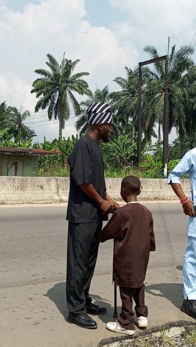 The image depicts a man and a young boy standing by the side of a road in Agburu Uke, Nigeria, under a bright, sunny sky with scattered white clouds. The man, who appears to be of African descent, is dressed in a black traditional two-piece garment and a black and white striped cap. He has a beard and is looking towards his right (viewer's left), holding the hand of the boy. The boy, also appearing to be of African descent, is shorter than the man and wears a brown traditional outfit. He faces away from the camera and is using a black walking stick, suggesting a mobility challenge. His short hair displays a textured pattern.
Partially visible on the far right is another individual in a light blue outfit, wearing a red bracelet. The background features lush tropical vegetation, including numerous tall palm trees and dense green foliage, possibly banana plants. A concrete barrier separates the road from the vegetated area. A small, rustic-looking green shack is visible behind the man, and a utility pole with wires stands further back on the right. The ground consists of asphalt road and a thin strip of dirt and grass. The scene portrays a moment of care or guidance as the man assists the boy, set against a vibrant tropical backdrop. There is no visible text.