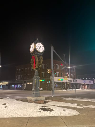This media file presents a tranquil, wintry street scene at night in Leominster, United States. The primary subject is a large, antique-style street clock with multiple faces, prominently adorned with a festive green wreath and a bright red bow, suggesting the holiday season. The clock faces feature Roman numerals, with the hands on one visible face indicating approximately 10:10.
The clock is mounted on a sturdy, decorative pole that also appears to support street lighting. In the background, across a visible road, stands a block of buildings, primarily constructed of brick, with numerous windows. A prominent green and yellow sign for "Subway" is clearly visible on one of these buildings, indicating a commercial establishment. Other less discernible signs, including one that might read "B&B" and "Leominster," can be seen further down the street. Traffic lights are also visible, contributing to the urban setting.
The ground, including the sidewalk and parts of the street, is covered in patches of snow and ice, indicating cold weather conditions. The sky is dark, affirming the nighttime setting, with artificial lights from the street clock, buildings, and traffic signals illuminating the scene. No people are visible, lending a quiet and still atmosphere to the urban landscape.