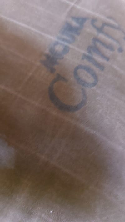 The image is a close-up, angled shot of a brown, textured surface, possibly fabric or paper, with dark blue lettering. The lettering is partially obscured and appears to be part of a brand name or logo. The visible words are "Ancira" and "Comfy." The texture of the surface suggests it might be a blanket, a tarp, or packaging material. There are diagonal lines in the background, which could be part of the surface texture or a separate layer. The lighting is uneven, with some areas brighter than others, suggesting a light source from the top left. The image lacks any discernible details about a specific location in New Nyanya, Nigeria, or any people or actions.