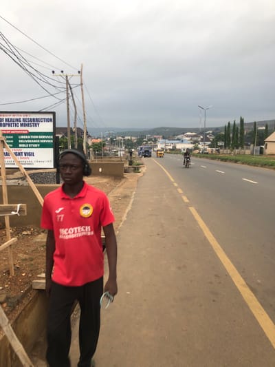 The image shows a man standing next to a roadside sign, beside a road with a yellow line. The sign advertises a "Healing Resurrection Prophetic Ministry" located in Karu, specifically referencing locations like Wangede and Jikwoyi FCT. The man is wearing a red shirt with "ECOTECH Conditioners" on it and the number 17. He holds something in his hand. In the background, there's a street scene with vehicles, buildings, and a cloudy sky. Power lines and poles are also visible.