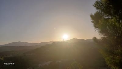 The image captures a sunset view in Benahavís, Spain, with a cluster of buildings visible on a hillside in the mid-ground, silhouetted against the bright sun. A prominent cross stands tall on the hill above the buildings, illuminated by the setting sun's rays. Rolling hills extend into the background, fading into a hazy mountain range under a pale, clear sky. In the foreground, the branches of a pine tree are visible on the right side of the frame. The bottom left corner displays the text "Galaxy A16." The overall scene is serene and bathed in the warm glow of the setting sun.