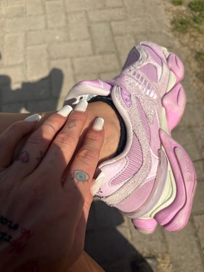 A close-up shot depicts a person's hand wearing a pink New Balance 9060 sneaker. The hand, adorned with long white nails and tattoos, rests on or near the shoe. The sneaker is in focus, with the background appearing to be an outdoor paved area. There is no information to link the image with the city of Werl, Germany.