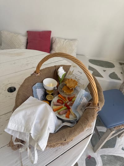 A woven basket containing a breakfast spread sits on a white wooden table. The basket is filled with a white cloth bag, small jars of jam, a salt shaker, a small white bowl of butter, a decorative napkin, a small glass dish with two mini quiches, a plate with sliced ham, cheese, cherry tomatoes, and carrot sticks, and a handwritten note. The table is set against a white wall with three pillows on a bench behind it. The floor is a mosaic of white and grey irregular shapes. In the background, a blue-cushioned chair is visible. The scene appears to be outdoors or in an open-air setting, possibly on the island of Naousa, Greece, given the architectural style of the floor and the overall aesthetic.