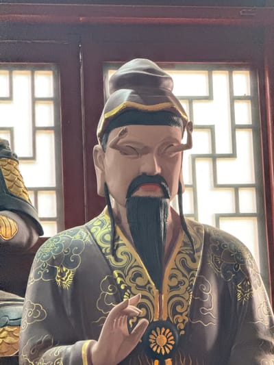 A statue of an ancient Chinese man, possibly a scholar or sage, with a long black beard and mustache, is dressed in a dark robe with gold embellishments. He wears a matching hat with gold trim. His head is bowed slightly, and his eyes are closed. His left hand is raised near his chest. The statue is located in Lintong District, China, as indicated by the architectural style of the latticed window in the background, which is common in traditional Chinese buildings in that area.
