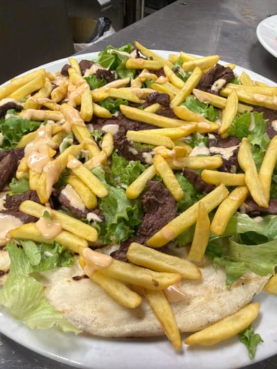 A close-up, overhead shot captures a dish of steak, fries, and salad served on a white plate, with a flatbread visible beneath the toppings. The steak is thinly sliced and scattered throughout the dish, interspersed with crinkle-cut french fries and pieces of crisp green lettuce. A creamy, pinkish sauce is drizzled over the entire dish. The plate is presented on a stainless steel countertop, suggesting a professional kitchen environment. The background is out of focus, but a blurred dark object and a hint of a yellow melon or squash are visible. The lighting is bright, casting minimal shadows. The scene is static, with no active subjects or interactions depicted. The location is identified as Parma, Italy.