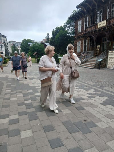 In Truskavets, Ukraine, two women are captured in a vertical photograph, standing on a paved walkway. One woman, in the foreground, wears a light-colored headscarf and a matching loose-fitting outfit with a cross-body bag. The other woman, positioned slightly behind and to her left, is also dressed in a similar light-colored, loose-fitting ensemble.

Behind them, a historic wooden building with intricate carvings and a sign reading "МУЗЕЙ" (Museum) in Cyrillic is visible, suggesting a cultural or historical site. The walkway is constructed with interlocking gray and beige pavers. In the background, trees and a cluster of buildings indicate a town or city setting. The sky appears overcast, suggesting a cloudy day. The overall mood seems relaxed and casual, possibly a sightseeing outing. Other individuals are partially visible in the background, also walking on the paved area.